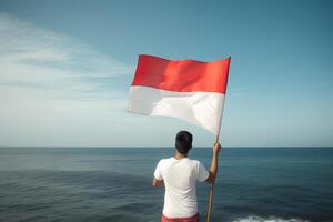 un' uomo Tenere un' rosso e bianca Indonesia bandiera guardare a il oceano. ai generato foto