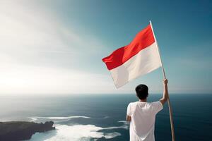 un' uomo Tenere un' rosso e bianca Indonesia bandiera guardare a il oceano. ai generato foto