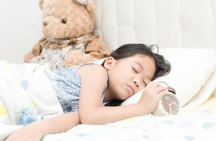 carino poco asiatico ragazza dormire e toccare allarme orologio su letto nel il Camera da letto. foto