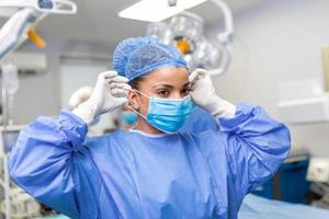 ritratto di bellissimo femmina medico chirurgo mettendo su medico maschera in piedi nel operazione camera. chirurgo a moderno operativo camera foto