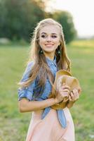 sorridente giovane donna detiene un' cappello nel sua mani mentre a piedi nel il estate all'aperto al di fuori il città su vacanza foto