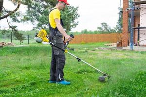 maschio giardiniere falcia il verde erba di il prato nel il Giardino dietro la casa a costruzione luogo con un' benzina tosaerba. trimmer per il cura di un' giardino tracciare foto