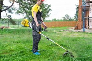 maschio giardiniere falcia il verde erba di il prato nel il Giardino dietro la casa a costruzione luogo con un' benzina tosaerba. trimmer per il cura di un' giardino tracciare foto