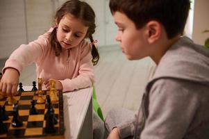 ritratto di un' concentrato 4 anni vecchio poco ragazza raccolta su un' scacchi pezzo mentre fabbricazione mossa su scacchi asse, giocando con sua fratello chi insegna sua il gioco foto