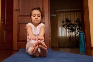 adorabile bambino ragazza fare allungamento esercizi, seduta a piedi nudi su Palestra stuoia mentre esercizio e praticante yoga in casa foto