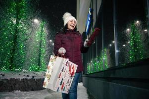 un' donna con presente scatola e shopping borse passeggiate giù il città strada illuminato di ghirlande, nel nevoso inverno sera foto