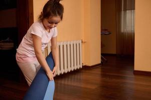 caucasico sorridente poco bambino ragazza nel abbigliamento sportivo, dispiegarsi blu fitness stuoia prima Lavorando su a casa. yoga la pratica. foto