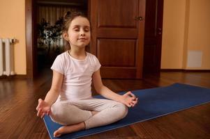 carino bambino praticante yoga al chiuso, si siede su fitness stuoia con sua occhi Chiuso, Meditare nel loto posa e mudra gesto foto