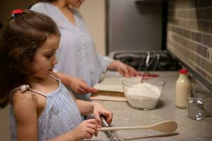 bellissimo bambino, adorabile poco ragazza in piedi a cucina contatore superiore, Tenere un' di legno cucchiaio su il sfocato sfondo di sua mamma, ricerca un' ricetta su ricetta libro per preparazione pancake Impasto foto