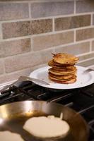 un' pila di appena al forno fatti in casa Pancakes su un' piatto Il prossimo per un' nero fornello. nel il sfocato primo piano è un' frittura padella con liquido Impasto Pancakes. Shrove martedì concetto foto