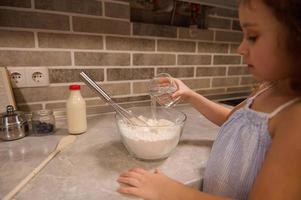 messa a fuoco su il mano di adorabile poco bambino ragazza, Tenere un' bicchiere con acqua e scrosciante esso in un' ciotola con Farina per preparazione liquido Impasto per Shrove Pancakes a casa cucina. foto