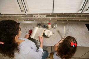 alto Visualizza di madre e figlia cucinando cibo insieme mentre in piedi a il cucina controsoffitto. mamma Tenere ricetta libro pronto per insegnare sua adorabile bambino ragazza Come per cucinare Impasto foto