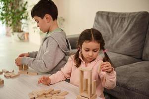 Due adorabile caucasico figli, concentrato bambini, ragazzo e ragazza giocando tavola Giochi, edificio di legno costruzioni su il tavolo a casa. educativo tempo libero e famiglia passatempo concetto foto