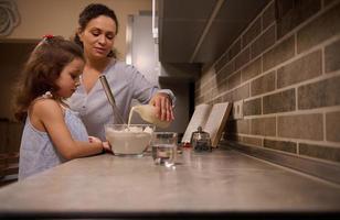 stile di vita ritratto di un' mamma scrosciante latte in ciotola di Farina e miscelazione ingredienti con un' frusta per preparare pancake Impasto. madre e figlia - bellissimo bambino siamo cucinando insieme nel il casa cucina foto