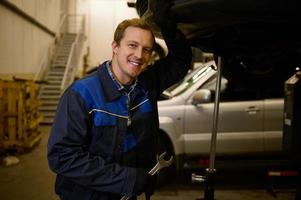 ritratto di un' giovane meccanico nel professionale uniforme, Tenere un' chiave inglese e Lavorando nel auto servizio con sollevato veicolo, sorrisi trentadue denti Sorridi lookng a telecamera. auto riparazione e Manutenzione. foto