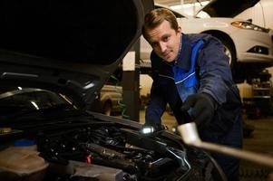 professionale meccanico, caucasico uomo nel complessivamente opera uniforme l'esame messa a punto, fissaggio, riparazione auto motore, veicolo parti utilizzando utensili attrezzatura nel laboratorio box auto. supporto e auto riparazione servizio foto