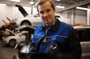 bello sorridente caucasico auto meccanico, tecnico nel uniforme, Tenere un' chiave inglese e in piedi contro sollevato macchine con Aperto cappuccio nel il riparazione negozio foto