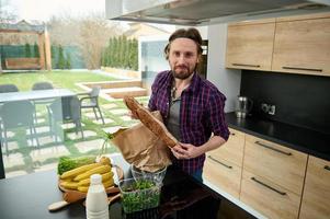 bello caucasico giovane uomo nel casuale Abiti detiene un' totale grano pane baguette, sembra a telecamera mentre disimballaggio il carta Borsa con salutare crudo cibo, verdure e frutta nel il casa cucina foto