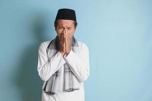 ritratto di attraente asiatico musulmano uomo nel bianca camicia starnuti perché di influenza, copertura il suo bocca con mano. isolato Immagine su blu sfondo foto