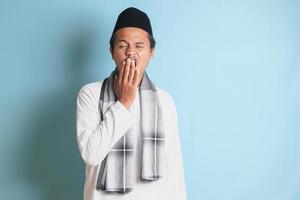 ritratto di attraente asiatico musulmano uomo nel bianca camicia sbadigli con mano copertura il suo bocca. isolato Immagine su blu sfondo foto