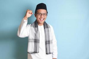 ritratto di asiatico musulmano uomo nel bianca koko camicia con zucchetto raccolta il suo cazzotto, festeggiare successo. isolato Immagine su grigio sfondo foto
