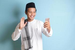 ritratto di asiatico musulmano uomo nel bianca koko camicia con zucchetto assunzione immagine di lui stesso o autoscatto, detto Ciao e agitando il suo mano durante video chiamata. isolato Immagine su blu sfondo foto
