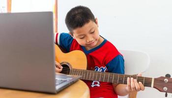 il storia di un' ragazzo Guardando un' taccuino computer mentre preparazione per pratica giocando chitarra a casa. ragazzi prendere classico chitarra Lezioni in linea. foto