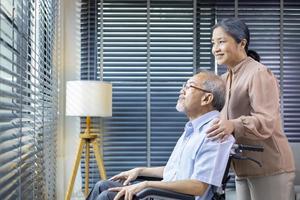 asiatico anziano uomo seduta nel sedia a rotelle a casa guardare su il finestra con il suo moglie è supporto e cura per amore e la pensione famiglia concetto foto