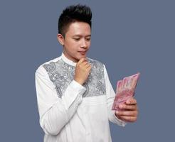 giovane uomo indossare koko camicia Tenere indonesiano uno centinaio mille rupia i soldi con dito su mento foto