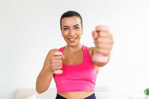 domestico formazione con pesi. positivo nero signora fare esercizi con manubri, rafforzamento sua corpo a casa. sorridente giovane femmina Lavorando su sua bicipite muscoli, soggiorno salutare foto