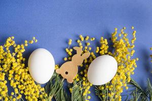 Pasqua sfondo, bianca uova e un' di legno lepre su un' blu sfondo, decorato con mimosa fiori, piatto, superiore Visualizza, vuoto spazio per testo. foto