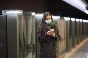 una donna con una maschera medica sta aspettando un treno e tiene in mano uno smartphone