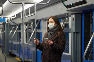 una donna con una maschera è in piedi e utilizza uno smartphone in un moderno vagone della metropolitana foto