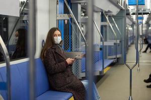 una donna con una maschera è seduta e utilizza uno smartphone in un moderno vagone della metropolitana foto