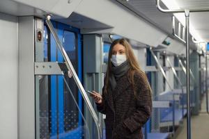 una donna con una maschera è in piedi e utilizza uno smartphone in un moderno vagone della metropolitana foto