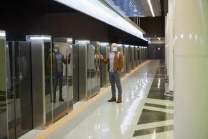 un uomo con una maschera facciale sta usando uno smartphone mentre aspetta un treno della metropolitana foto