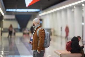 un uomo con una maschera facciale sta usando uno smartphone mentre aspetta un treno della metropolitana foto