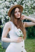 bellissimo giovane ragazza nel bianca vestito e cappello nel fioritura Mela frutteto. fioritura Mela alberi con bianca fiori. foto