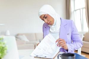 giovane Arabia attività commerciale donna Lavorando nel a casa, puntamento su computer portatile, copia spazio. musulmano femmina Lavorando con computer nel il camera , scrittura carta. giovane musulmano donna utilizzando il computer portatile foto