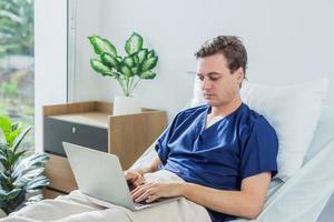 caucasico paziente utilizzando il computer portatile nel ospedale camera. uomo malato paziente dire bugie nel un' ospedale letto foto