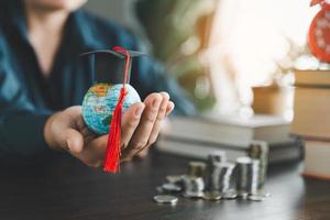 mano Tenere la laurea berretto su globo con pila moneta. concetto di Salvataggio i soldi per un' Borsa di studio per studia all'estero a un' Università livello. finanziario pianificazione contabilità idee per futuro formazione scolastica. foto