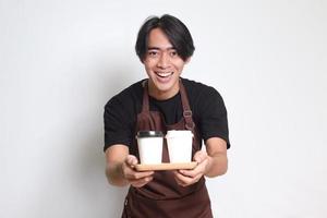 ritratto di attraente asiatico barista uomo nel Marrone grembiule Tenere Due prendere lontano carta caffè tazze. isolato Immagine su bianca sfondo foto