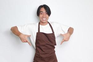 ritratto di attraente asiatico barista uomo nel Marrone grembiule mostrando Prodotto, puntamento a qualcosa con mani. pubblicità concetto. isolato Immagine su bianca sfondo foto