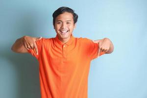 eccitato asiatico uomo nel arancia camicia sorridente e guardare a il telecamera puntamento con Due mani e dita per il lato. isolato Immagine su blu sfondo foto