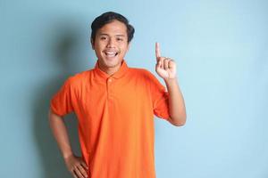 allegro asiatico uomo nel arancia camicia in piedi contro blu sfondo, puntamento su il suo indice e ricordando qualcosa per fare foto