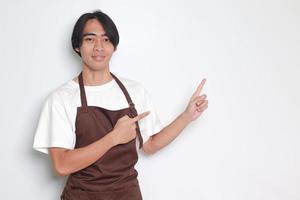 ritratto di attraente asiatico barista uomo nel Marrone grembiule mostrando Prodotto, puntamento a qualcosa con mani. pubblicità concetto. isolato Immagine su bianca sfondo foto