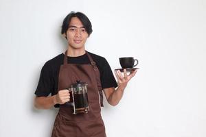 ritratto di attraente asiatico barista uomo nel Marrone grembiule mostrando un' tazza e piattino mentre Tenere francese stampa caffè creatore. isolato Immagine su bianca sfondo foto