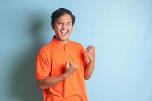 ritratto di eccitato asiatico uomo nel arancia camicia raccolta il suo cazzotto, festeggiare successo. isolato Immagine su blu sfondo foto