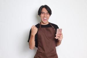 ritratto di attraente asiatico barista uomo nel Marrone grembiule Tenere mobile Telefono e Guardando video mentre raccolta il suo cazzotto festeggiare successo. isolato Immagine su bianca sfondo foto