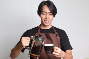 ritratto di attraente asiatico barista uomo nel Marrone grembiule scrosciante caffè in un' tazza a partire dal francese stampa caffè creatore. isolato Immagine su bianca sfondo foto